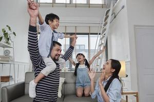 Asian Thai family together, dad plays and teases with children and mum by carrying and holding son on shoulders in home living room, happy leisure times, lovely weekend, wellbeing domestic lifestyle. photo