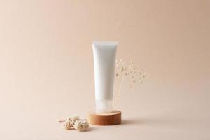 White tube of cosmetic cream with flowers on wooden podium on beige background. Close up. photo