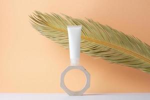 White tube of cosmetic cream with palm leaf on wooden podium on beige background. Close up. photo