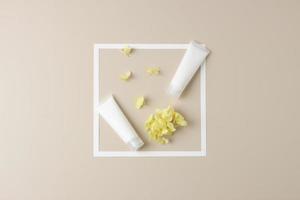 White tubes of cosmetic cream with white frame and flowers on pastel beige background. Flat lay, copy space photo