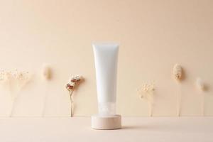 White tube of cosmetic cream with flowers on wooden podium on beige background. Close up. photo