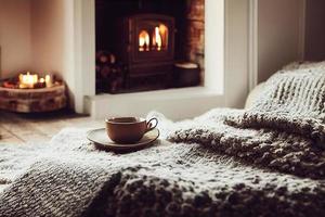 Mug with hot tea standing on a chair with woolen blanket in a cozy living room with fireplace photo