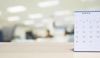 White paper desk calendar on wood table top with blurred office interior background photo
