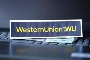 TERNOPIL, UKRAINE - SEPTEMBER 6, 2022 Western Union paper logotype lies on black laptop with US dollar bills. Western Union Company is American multinational financial services company photo