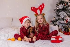 dos niños en casa en pijama para navidad están clasificando regalos y acurrucándose en la cama contra el fondo de un árbol de navidad foto