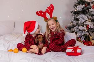 dos niños en casa en pijama para navidad están clasificando regalos y acurrucándose en la cama contra el fondo de un árbol de navidad foto