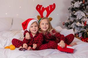 dos niños en casa en pijama para navidad están clasificando regalos y acurrucándose en la cama contra el fondo de un árbol de navidad foto