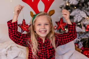 niña rubia feliz en casa en pijama para Navidad clasificando regalos y jugando con juguetes de Navidad foto