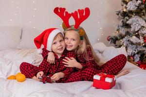 dos niños en casa en pijama para navidad están clasificando regalos y acurrucándose en la cama contra el fondo de un árbol de navidad foto