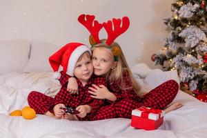 dos niños en casa en pijama para navidad están clasificando regalos y acurrucándose en la cama contra el fondo de un árbol de navidad foto