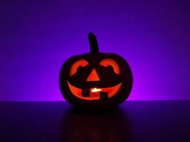 Carved glowing pumpkin jack-o-lantern with candle inside stands on surface in dark night and blurred defocused purple background. The back wall highlighted in violet. Halloween holiday concept. photo