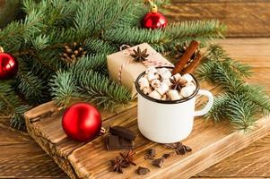 white enameled mug with hot chocolate or cocoa with marshmallows on the wooden pedium against the background of a green branch of spruce.
