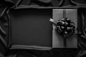 A black gift box with ribbon puts on black smooth and wavy cloth with space for text. Black friday and Boxing day concept. photo