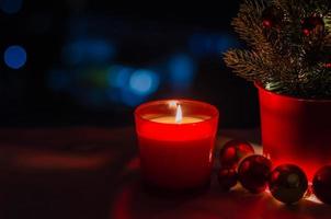 Candle with flame in red glass and Christmas tree decorated with bauble ornaments on dark background. photo