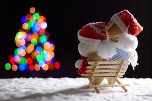 Brown and white teddy bear wearing santa hat sitting on wooden bench when snowing in winter looking at colorful bokeh lights of Christmas tree. photo