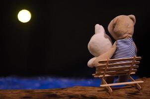 muñecos de oso de peluche y conejo actuando como pareja sentados en un banco hecho de palitos de helado y mirando la luna en la noche romántica en la orilla del mar azul. foto