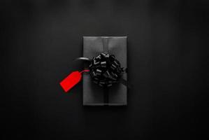 A black gift box with red ribbon and blank price tag puts on black background. Black friday and Boxing day concept. photo