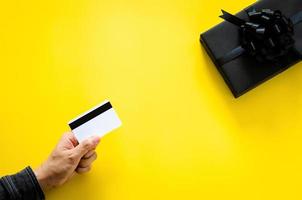 mano con tarjeta de crédito para comprar una caja de regalo negra sobre fondo amarillo para el concepto de viernes negro y lunes cibernético. foto