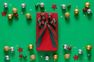 Red gift box with bow ribbon and Christmas baubles ornaments on green background. Christmas and New year concept. photo