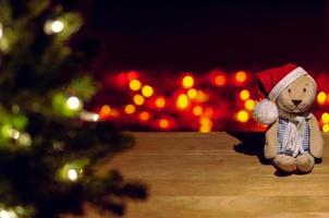 Santa Claus teddy bear sitting on wooden table with Christmas tree and colorful bokeh lights background. photo