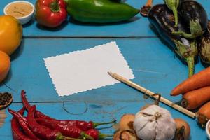 Vegetables are laid out around a sheet of paper and a pencil. Empty space for text. Vegetables, empty blank for recipe on a blue background. photo