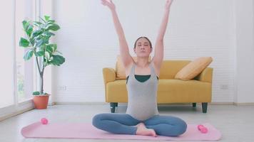 Beautiful young Asian pregnant woman in sportswear is doing yoga at home. video