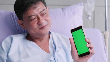 älterer asiatischer männlicher patient, der während des krankenhausaufenthaltes ein smartphone mit grünem bildschirm hält. video