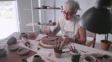 Aziatisch ouderen vrouw genieten van pottenbakkerij werk Bij huis. een vrouw keramist is maken nieuw pottenbakkerij in een studio video