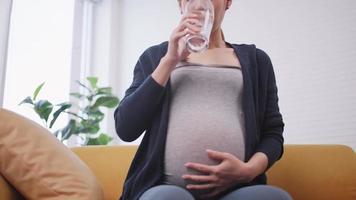 mujer asiática embarazada bebiendo agua en el sofá de casa. video