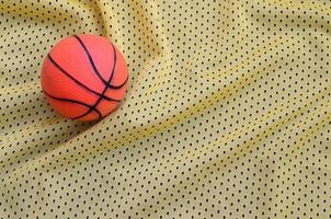 el pequeño baloncesto de goma naranja se encuentra sobre una textura de tela de jersey deportivo amarillo y un fondo con muchos pliegues foto