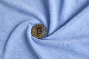 The golden bitcoin lies on a blanket made of soft and fluffy light blue fleece fabric with a large number of relief folds. The shape of the folds resembles a fan from a video card cooler photo