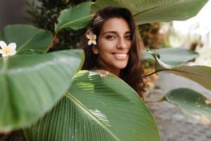 Young woman covers herself with huge green leaf and looks into camera with smile photo