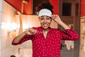 Curly woman in polka dot shirt and a sleep mask poses with a toothbrush in the bathroom. photo