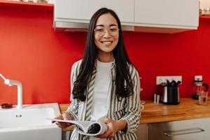 mujer de ojos marrones con gafas con una sonrisa mira a la cámara en la cocina y lleva un diario foto