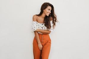 Picture of lady in orange trousers with high waist and white top with floral print. Brunette girl i