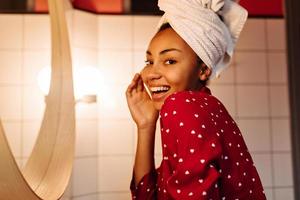 Brown-eyed woman in silk pajamas and towel on her head with smile looking at camera in bathroom.