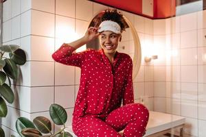 Pleased woman in polka-dot pajamas takes off her sleep mask and poses in bathroom with smile. photo