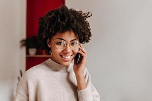 Curly woman in glasses smiling, looking at camera and talking on phone.