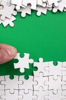 The hand folds a white jigsaw puzzle and a pile of uncombed puzzle pieces lies against the background of the green surface. Texture photo with space for text