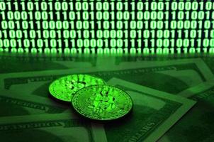 Two bitcoins lies on a pile of dollar bills on the background of a monitor depicting a binary code of bright green zeros and one units on a black background. Low key lighting photo