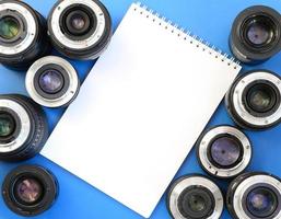 varias lentes fotográficas y un cuaderno blanco yacen sobre un fondo azul brillante. espacio para texto foto