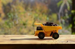 A small yellow toy truck is loaded with brown coffee beans. A car on a wooden surface against a background of autumn forest. Extraction and transportation of coffee photo