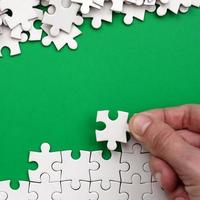 The hand folds a white jigsaw puzzle and a pile of uncombed puzzle pieces lies against the background of the green surface. Texture photo with space for text