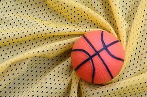 Small orange rubber basketball lies on a yellow sport jersey clothing fabric texture and background with many folds photo