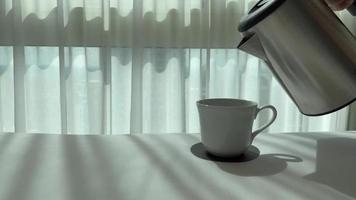 Hot water from the kettle pouring to a cup of coffee that put on white bed sheet with shadow of curtain in the morning. video