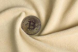 The golden bitcoin lies on a blanket made of soft and fluffy light orange fleece fabric with a large number of relief folds. The shape of the folds resembles a fan from a video card cooler photo