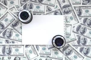 dos lentes fotográficos y un cuaderno blanco se encuentran en el fondo de muchos billetes de dólar. espacio para texto foto