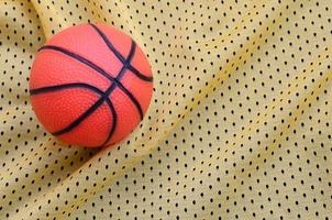 Small orange rubber basketball lies on a yellow sport jersey clothing fabric texture and background with many folds photo