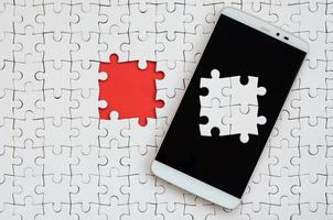 A modern big smartphone with several puzzle elements on the touch screen lies on a white jigsaw puzzle in an assembled state with missing elements photo