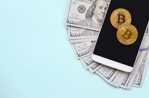 Bitcoins lies on a smartphone and hundred dollar bills on a light blue background photo